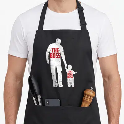 Father boss apron - White silhouettes of man and child holding hands with red distressed text reading THE BOSS THE REAL — shown on a black cotton canvas apron against a white background