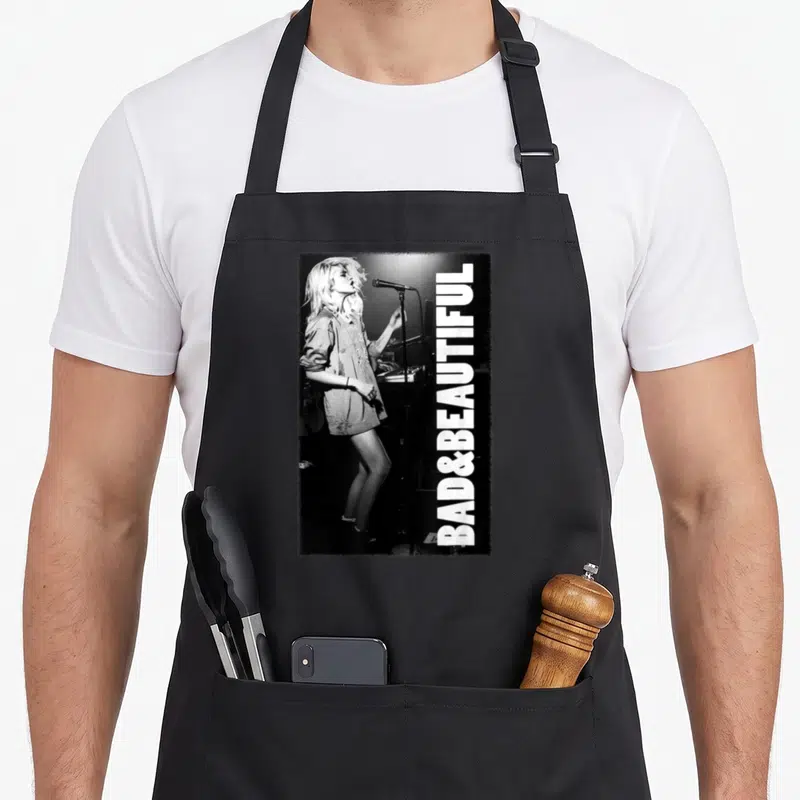 Inscription beautiful apron - Vertical BAD&BEAUTIFUL text beside grayscale photo of woman singing into microphone — shown on a black cotton canvas apron against a white background