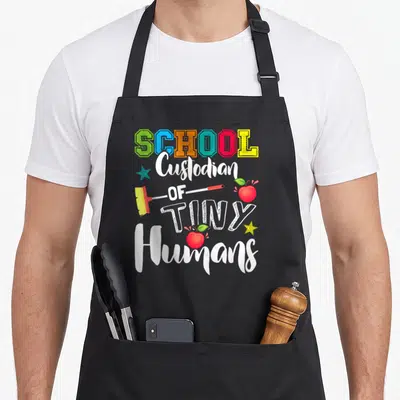 Tiny janitor apron - Stacked colorful text reading SCHOOL Custodian OF TINY Humans with broom, apples, and stars — shown on a black cotton canvas apron against a white background