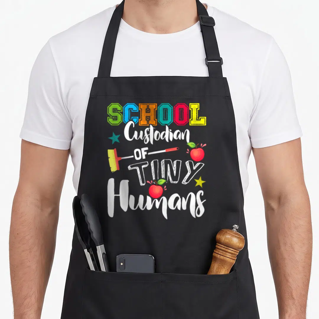 Tiny janitor apron - Stacked colorful text reading SCHOOL Custodian OF TINY Humans with broom, apples, and stars — shown on a black cotton canvas apron against a white background