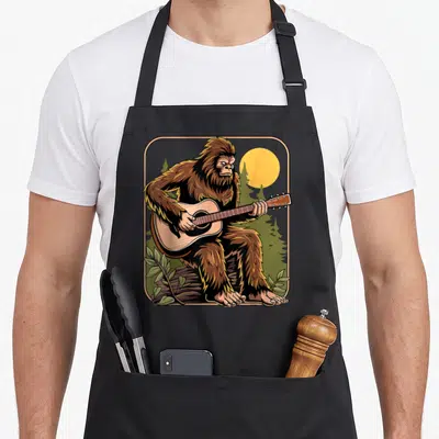 Guitarist bigfoot apron - Cartoon Bigfoot sitting on a log playing an acoustic guitar surrounded by a yellow sun — shown on a black cotton canvas apron against a white background