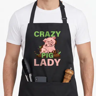 Pig agriculture farm apron - CRAZY PIG LADY text surrounding pink cartoon pig sitting in brown mud with green grass — shown on a black cotton canvas apron against a white background