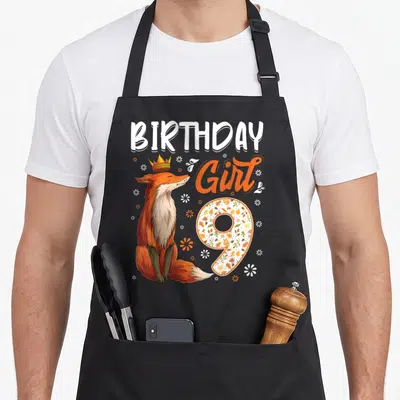 9th fox aprons - Crowned orange fox sitting beside BIRTHDAY Girl 9 text filled with autumn leaves and flowers — shown on a black cotton canvas apron against a white background