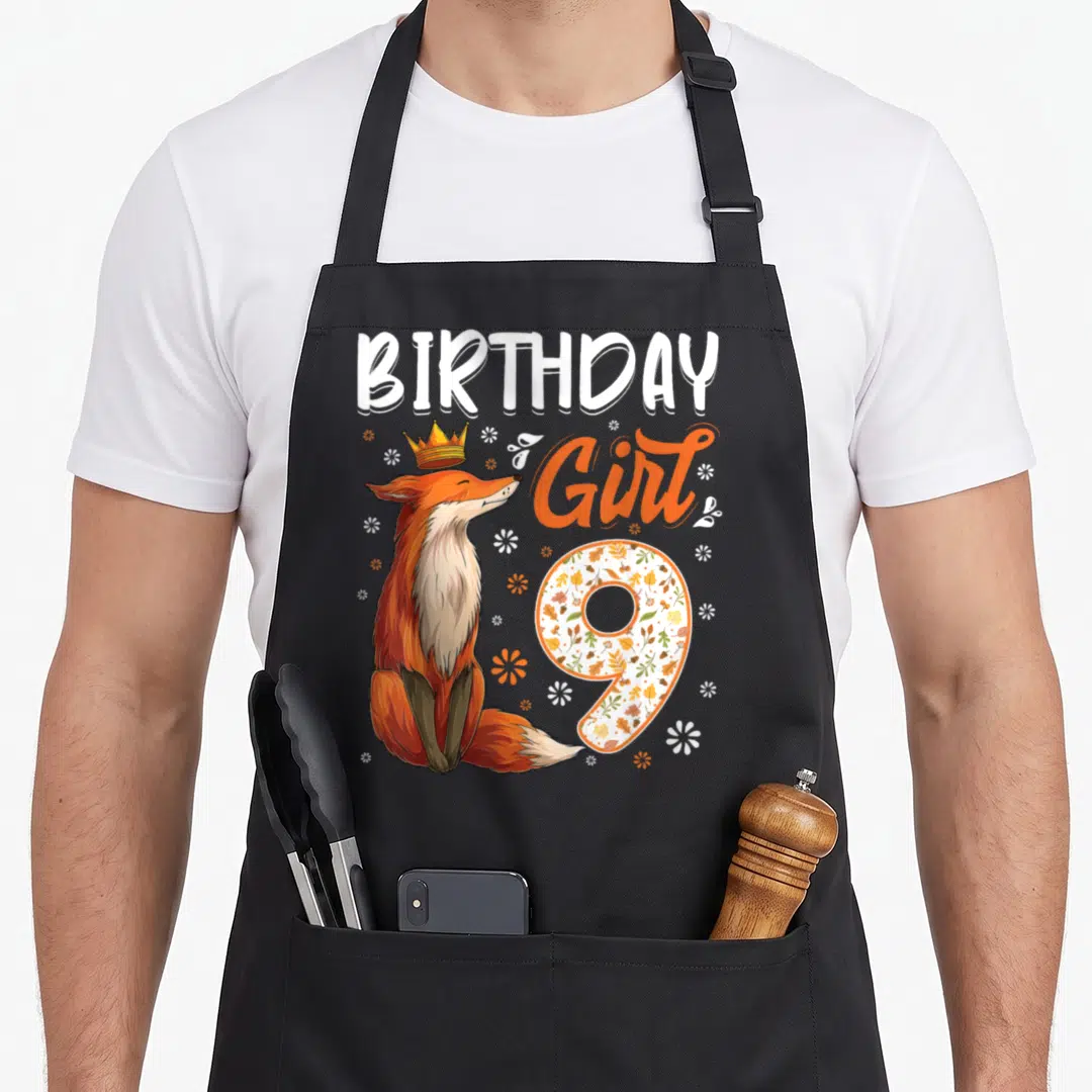 9th fox aprons - Crowned orange fox sitting beside BIRTHDAY Girl 9 text filled with autumn leaves and flowers — shown on a black cotton canvas apron against a white background