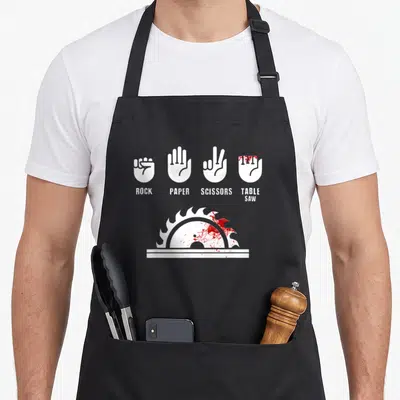 Carpenter paper apron - White rock paper scissors table saw hands with red blood splatters on saw blade — shown on a black cotton canvas apron against a white background