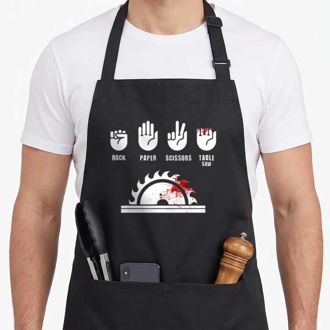 Carpenter paper apron - White rock paper scissors table saw hands with red blood splatters on saw blade — shown on a black cotton canvas apron against a white background