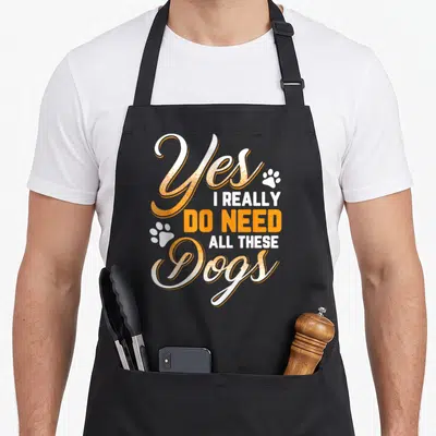 Funny these do i need an apron - Stacked orange and white text reading Yes I Really Do Need All These Dogs with two paw — shown on a black cotton canvas apron against a white background