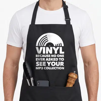 Record disc vinyl apron - White half-circle vinyl record graphic above bold stacked text reading VINYL and MP3 — shown on a black cotton canvas apron against a white background