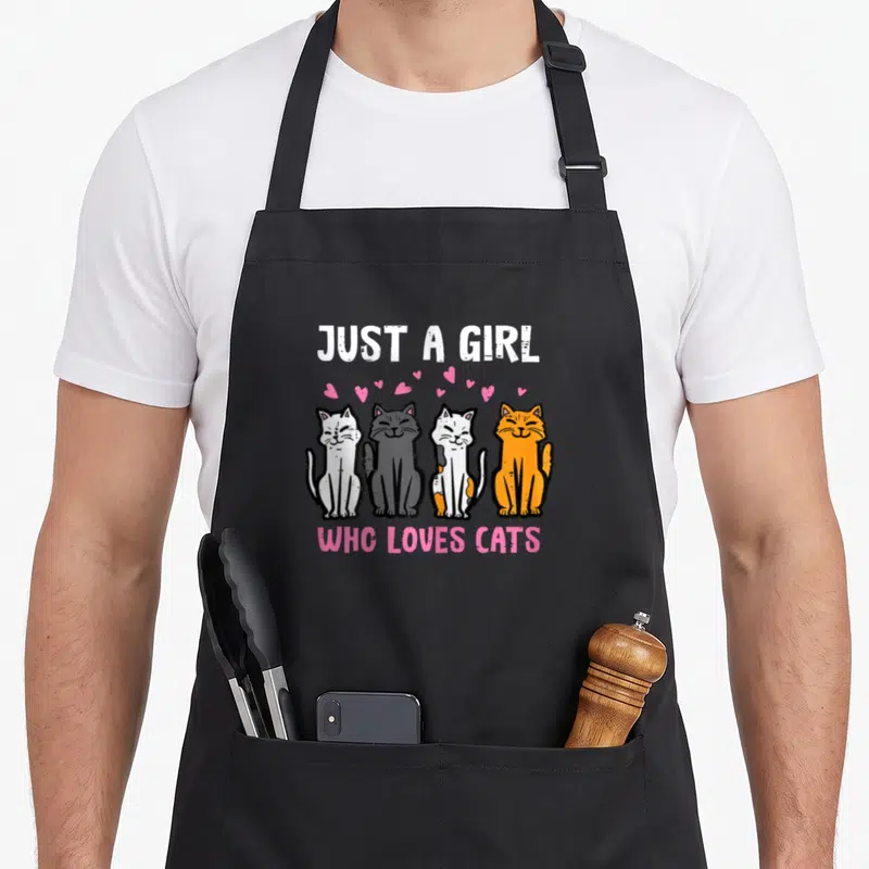 Youth cat apron - Four sitting cats in white, grey, and orange below pink hearts and text JUST A GIRL WHO LOVES CATS — shown on a black cotton canvas apron against a white background