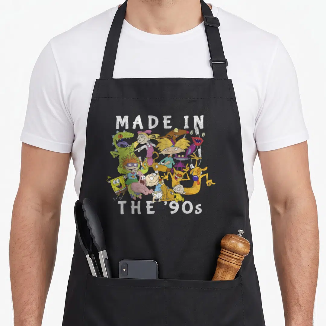 Faded character aprons - MADE IN THE '90s text with cartoon character cluster in green yellow pink purple white — shown on a black cotton canvas apron against a white background
