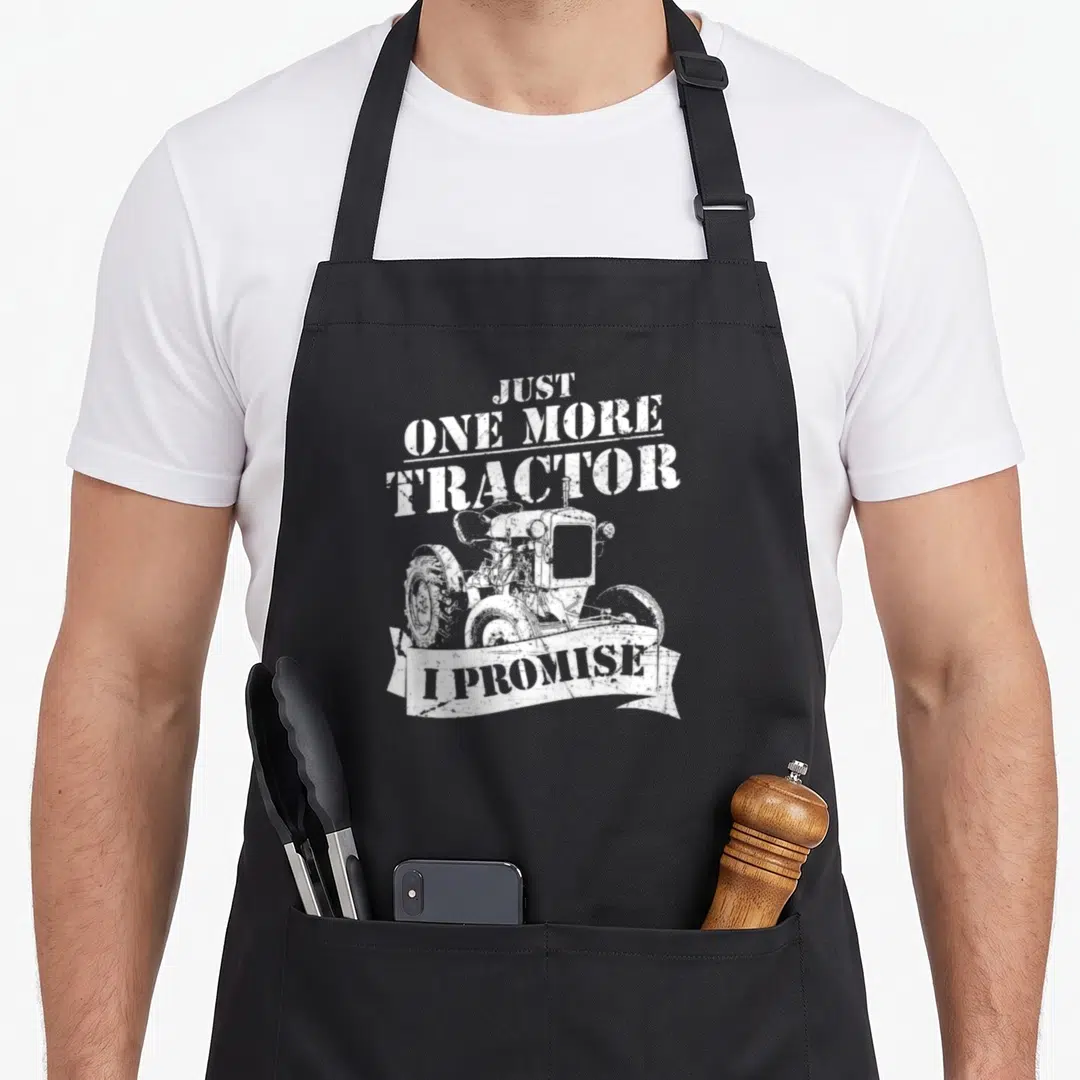 Cute tractor farmer apron - White distressed tractor illustration with text JUST ONE MORE TRACTOR I PROMISE in a ribbon — shown on a black cotton canvas apron against a white background