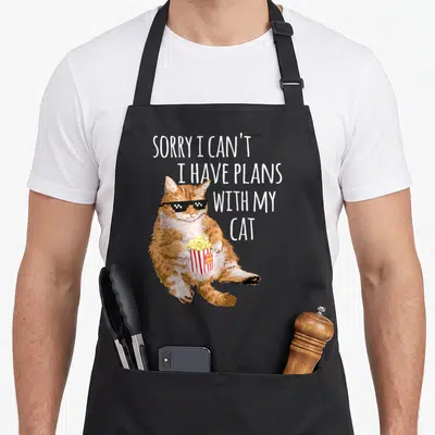 Sorry plan cat apron - Orange cartoon cat wearing pixel sunglasses holding popcorn bucket with stacked white text — shown on a black cotton canvas apron against a white background