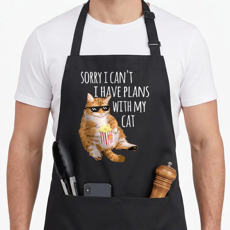 Sorry plan cat apron - Orange cartoon cat wearing pixel sunglasses holding popcorn bucket with stacked white text — shown on a black cotton canvas apron against a white background