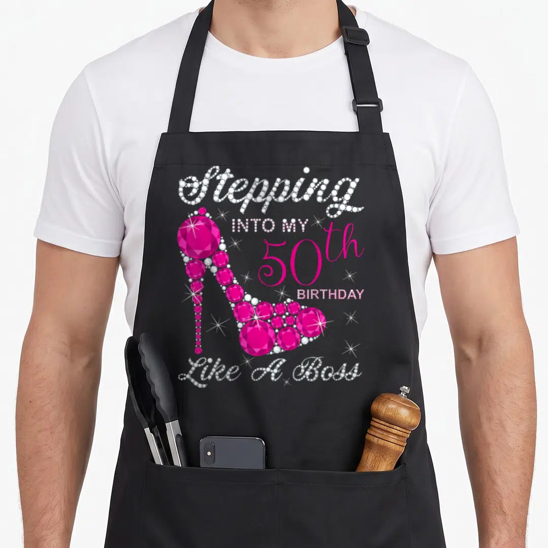Queen stepping bbq boss apron - Pink and white gem high heel shoe with Stepping Into My 50th Birthday Like A Boss text — shown on a black cotton canvas apron against a white background