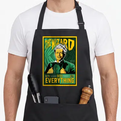 Wizard wicked apron - The Wizard Sees Everything text and man portrait in green and yellow with two eye symbols — shown on a black cotton canvas apron against a white background