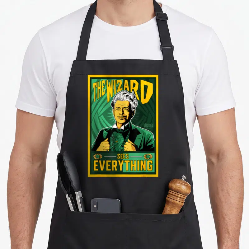 Wizard wicked apron - The Wizard Sees Everything text and man portrait in green and yellow with two eye symbols — shown on a black cotton canvas apron against a white background