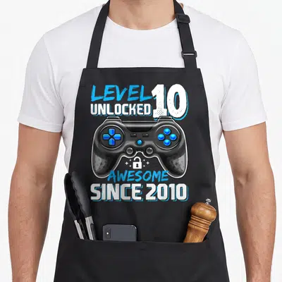 Birthday age video game apron - Black game controller with blue buttons and text Level 10 Unlocked Awesome Since 2010 — shown on a black cotton canvas apron against a white background