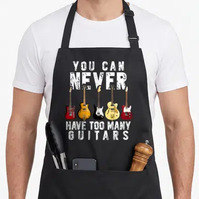 Many playing guitar apron - Five electric guitars in red, yellow, black, and cream below distressed white text reading — shown on a black cotton canvas apron against a white background