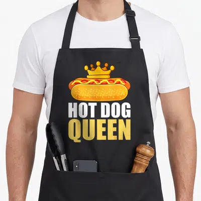 Grilled hot dog apron - Cartoon hot dog with crown and stacked HOT DOG QUEEN text in yellow and white — shown on a black cotton canvas apron against a white background