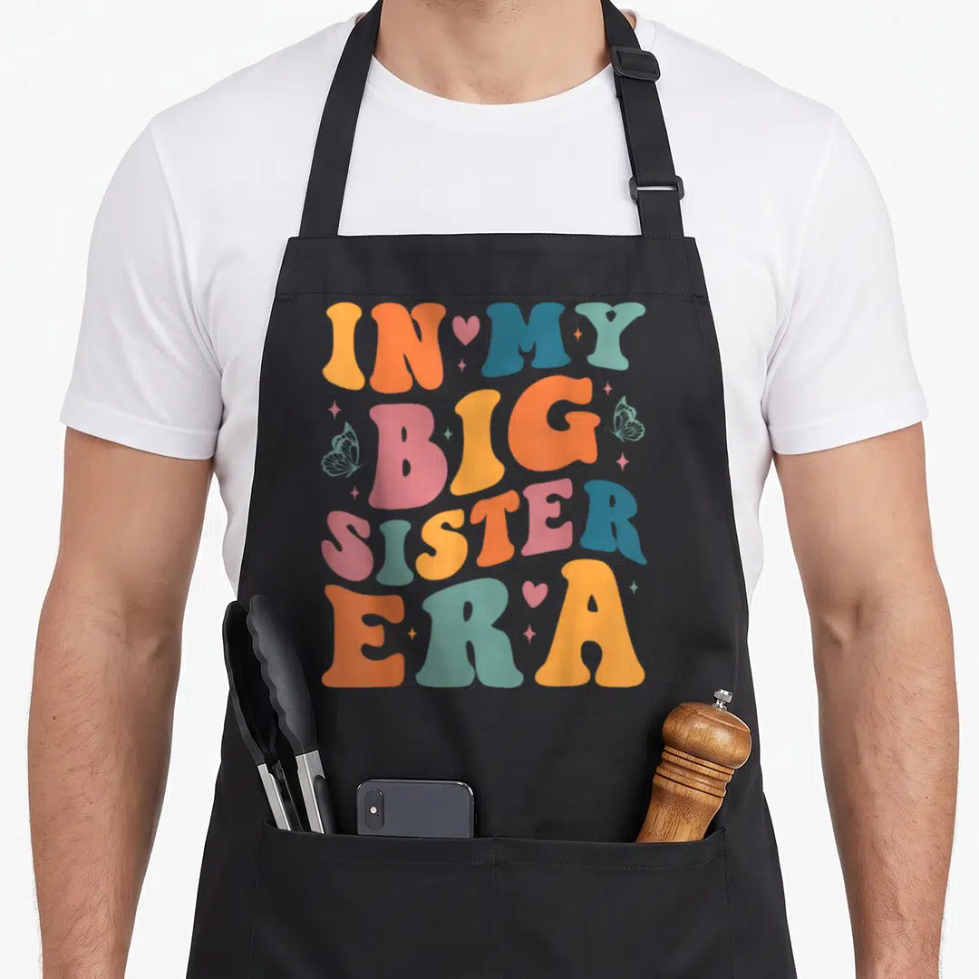 Era pregnancy announcement apron - IN MY BIG SISTER ERA text in orange pink blue teal yellow with hearts butterflies — shown on a black cotton canvas apron against a white background