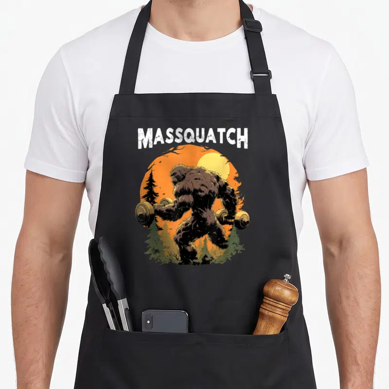 Hairy animal apron - MASSQUATCH text above brown Bigfoot holding a barbell against orange sun and green trees — shown on a black cotton canvas apron against a white background