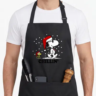 Snowfall holiday apron - Snoopy and Woodstock in red hats holding candy canes in snow with chillin text — shown on a black cotton canvas apron against a white background
