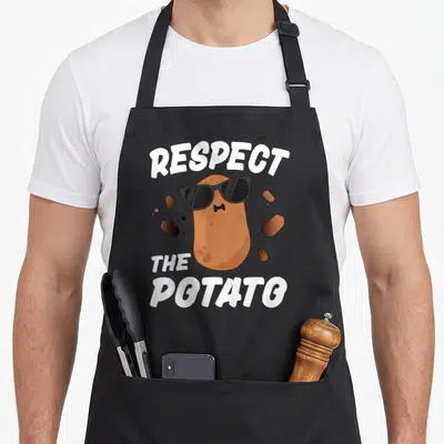 Respect potato vegetable apron - Brown cartoon potato wearing black sunglasses between white text reading Respect — shown on a black cotton canvas apron against a white background