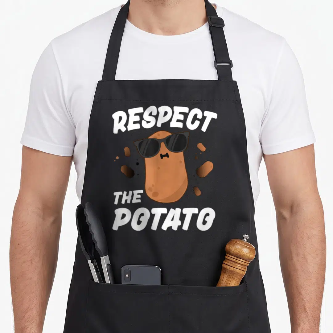 Respect potato vegetable apron - Brown cartoon potato wearing black sunglasses between white text reading Respect — shown on a black cotton canvas apron against a white background