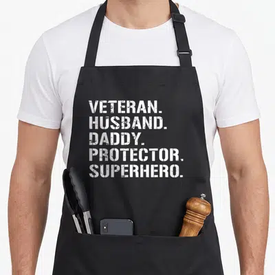Superhero best husband apron - White stenciled text stacked vertically reading VETERAN HUSBAND DADDY PROTECTOR SUPERHERO — shown on a black cotton canvas apron against a white background