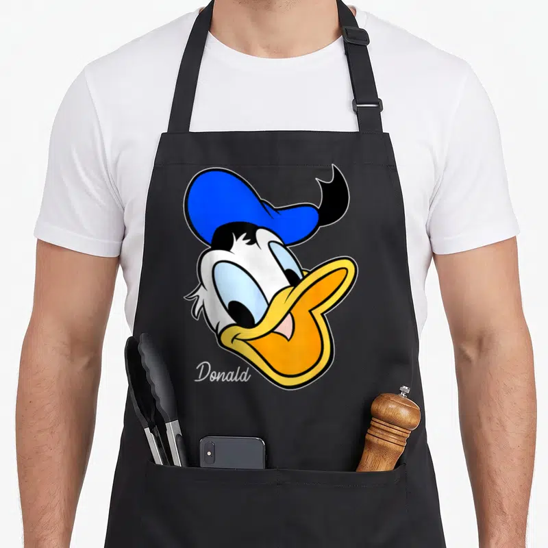 Face duck apron - Large Donald Duck head in blue yellow white and black with Donald text below — shown on a black cotton canvas apron against a white background
