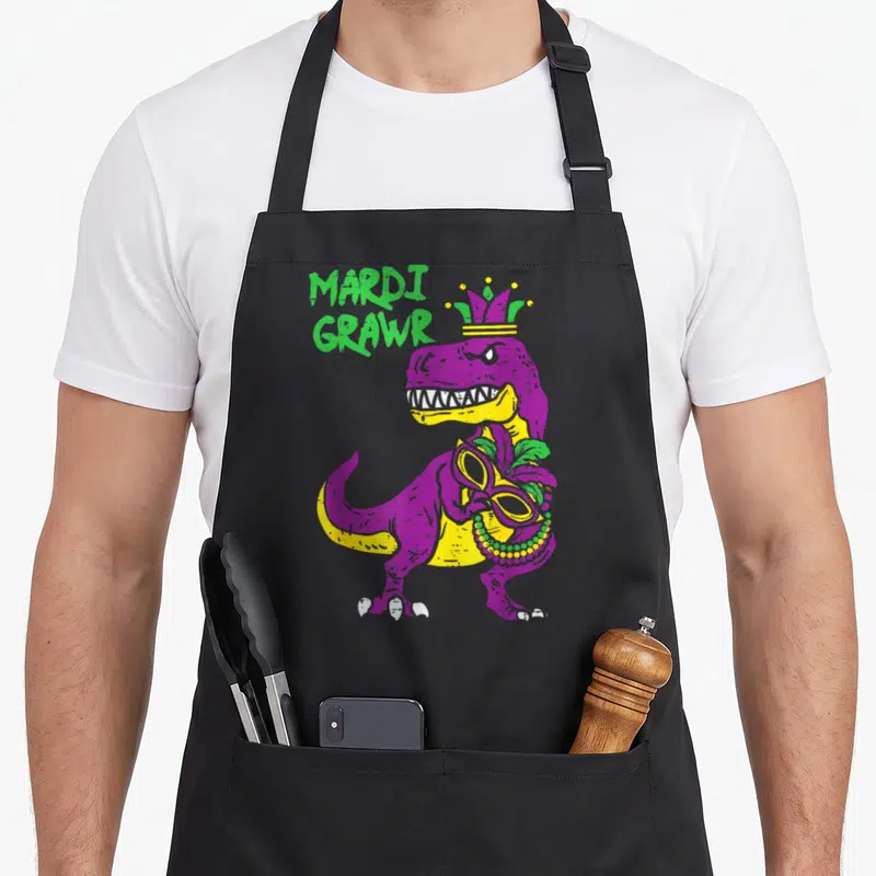 Rex mardi gras aprons - Purple and yellow T-Rex holding a beaded masquerade mask with green MARDI GRAWR text — shown on a black cotton canvas apron against a white background
