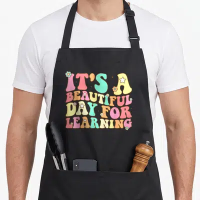 Beautiful teacher apron - Retro text IT'S A BEAUTIFUL DAY FOR LEARNING in pink yellow teal with stars and flowers — shown on a black cotton canvas apron against a white background