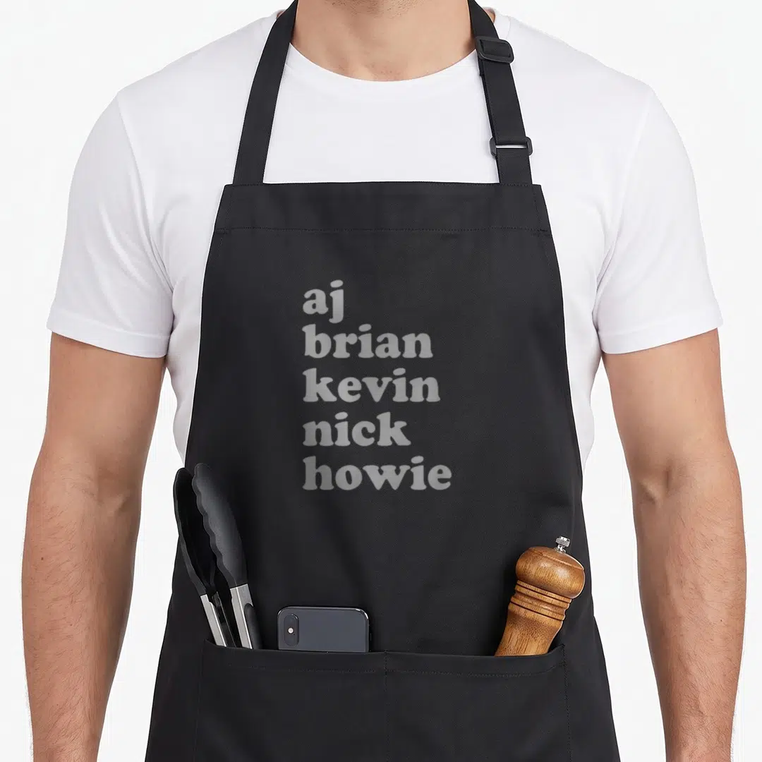 Stacked backstreet boys apron - White stacked text aj brian kevin nick howie on black background — shown on a black cotton canvas apron against a white background