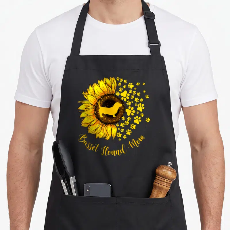 Basset mother's day aprons - Yellow sunflower with basset hound silhouette and paw prints arched text below — shown on a black cotton canvas apron against a white background
