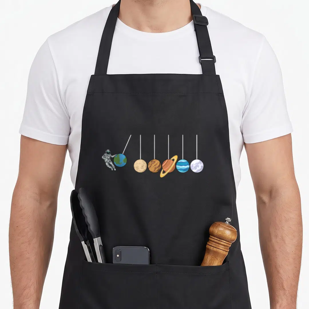 Solar astrophysicist apron - Cartoon astronaut holding Earth next to six planets hanging from white strings — shown on a black cotton canvas apron against a white background