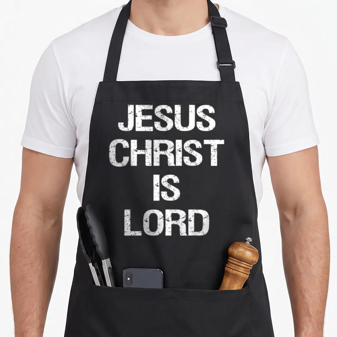 Jesus christ christian apron - White distressed text reading JESUS CHRIST IS LORD stacked in four lines — shown on a black cotton canvas apron against a white background