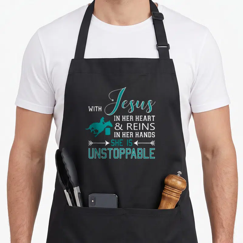 Jesus she in her apron - Teal silhouette of woman riding horse past barrel next to stacked white and teal typography — shown on a black cotton canvas apron against a white background
