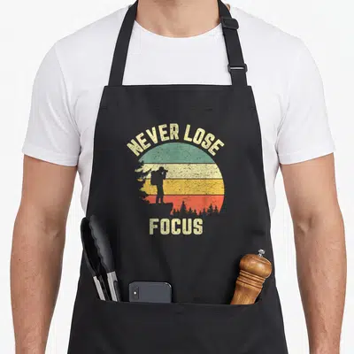 Lose photographer apron - Retro sun with colored stripes, photographer silhouette, and text NEVER LOSE FOCUS — shown on a black cotton canvas apron against a white background
