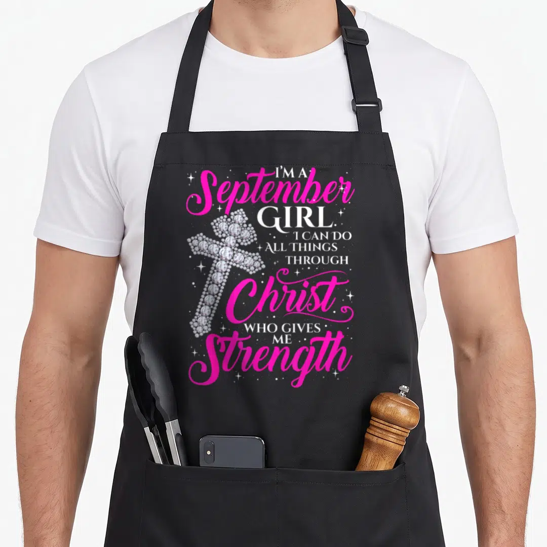 September christian apron - Diamond cross with pink and white text reading I'M A September GIRL I CAN DO ALL THINGS — shown on a black cotton canvas apron against a white background