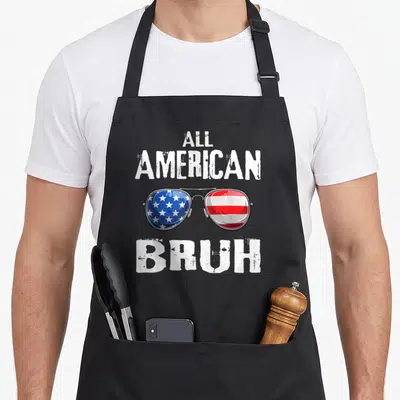 Bruh patriotic 4th of july apron - ALL AMERICAN BRUH text above and below sunglasses with star-spangled and striped — shown on a black cotton canvas apron against a white background