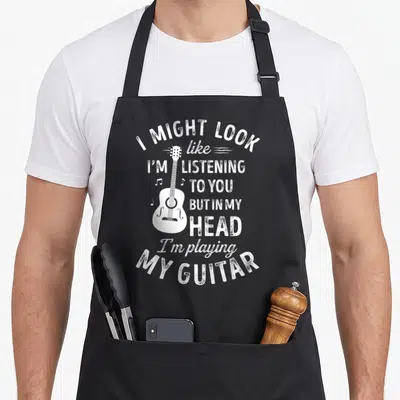 Might songwriter apron - White distressed acoustic guitar with text I might look like I'm listening to you but in my — shown on a black cotton canvas apron against a white background