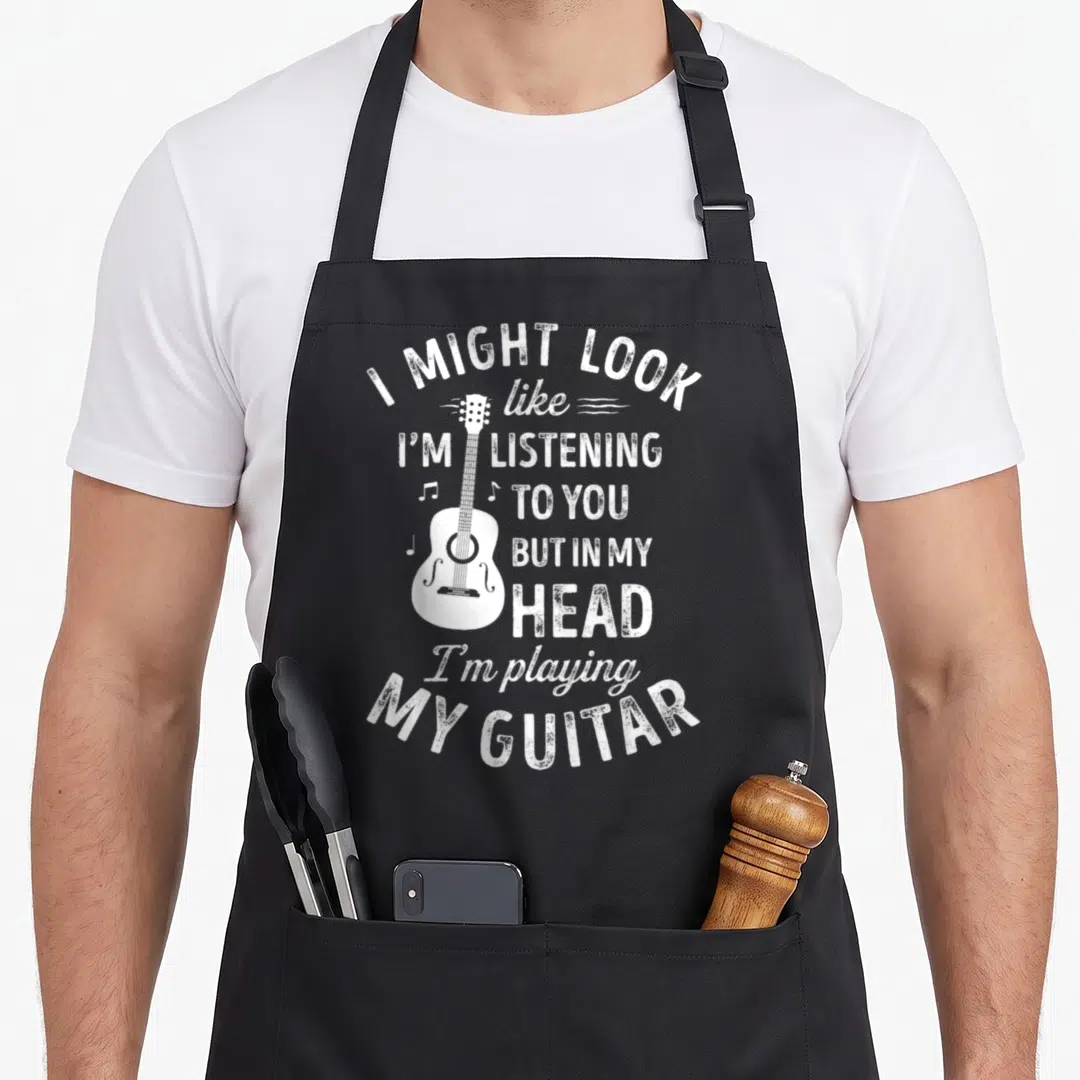 Might songwriter apron - White distressed acoustic guitar with text I might look like I'm listening to you but in my — shown on a black cotton canvas apron against a white background