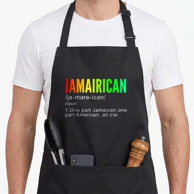 Jamairican jamaican apron - JAMAIRICAN text in red yellow green gradient with white definition below — shown on a black cotton canvas apron against a white background