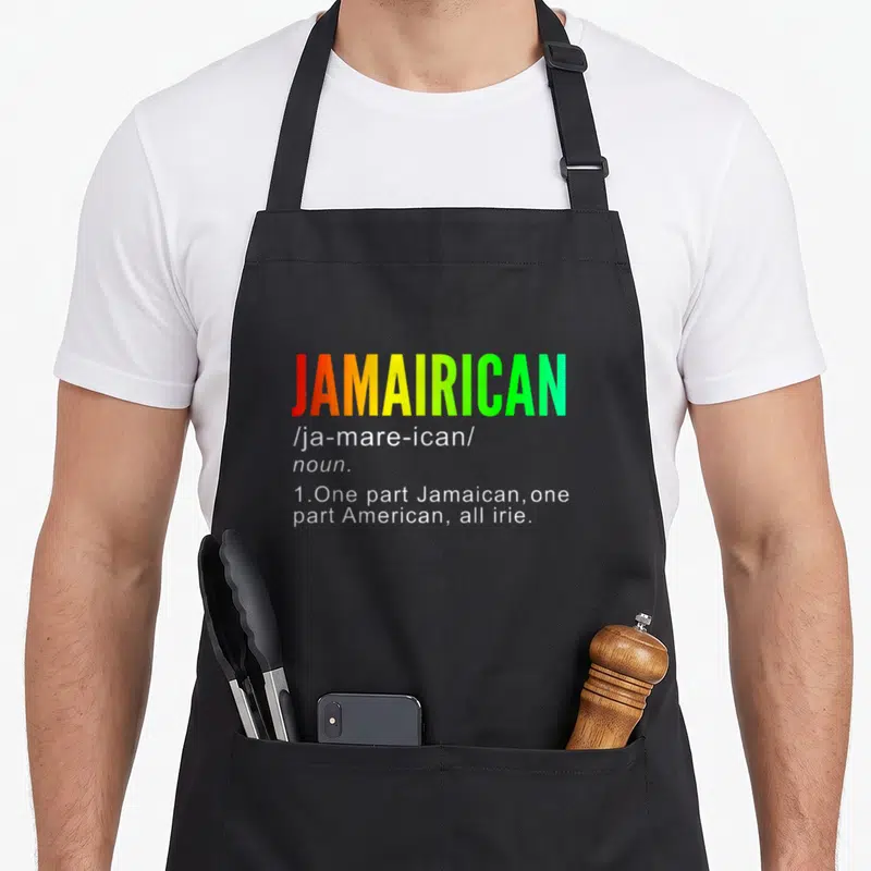 Jamairican jamaican apron - JAMAIRICAN text in red yellow green gradient with white definition below — shown on a black cotton canvas apron against a white background
