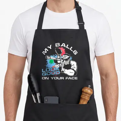 Ball paint apron - Paintball player holding marker with text MY BALLS LOOK GOOD ON YOUR FACE and splatters — shown on a black cotton canvas apron against a white background
