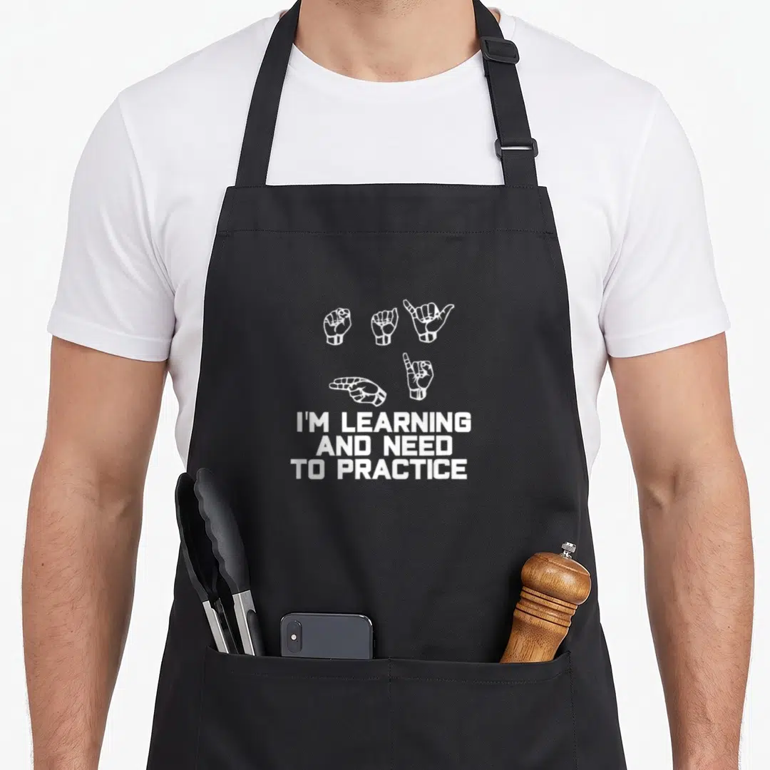 Learning asl sign for apron - Five white line-art hand gestures above three lines of text reading I'M LEARNING AND NEED — shown on a black cotton canvas apron against a white background