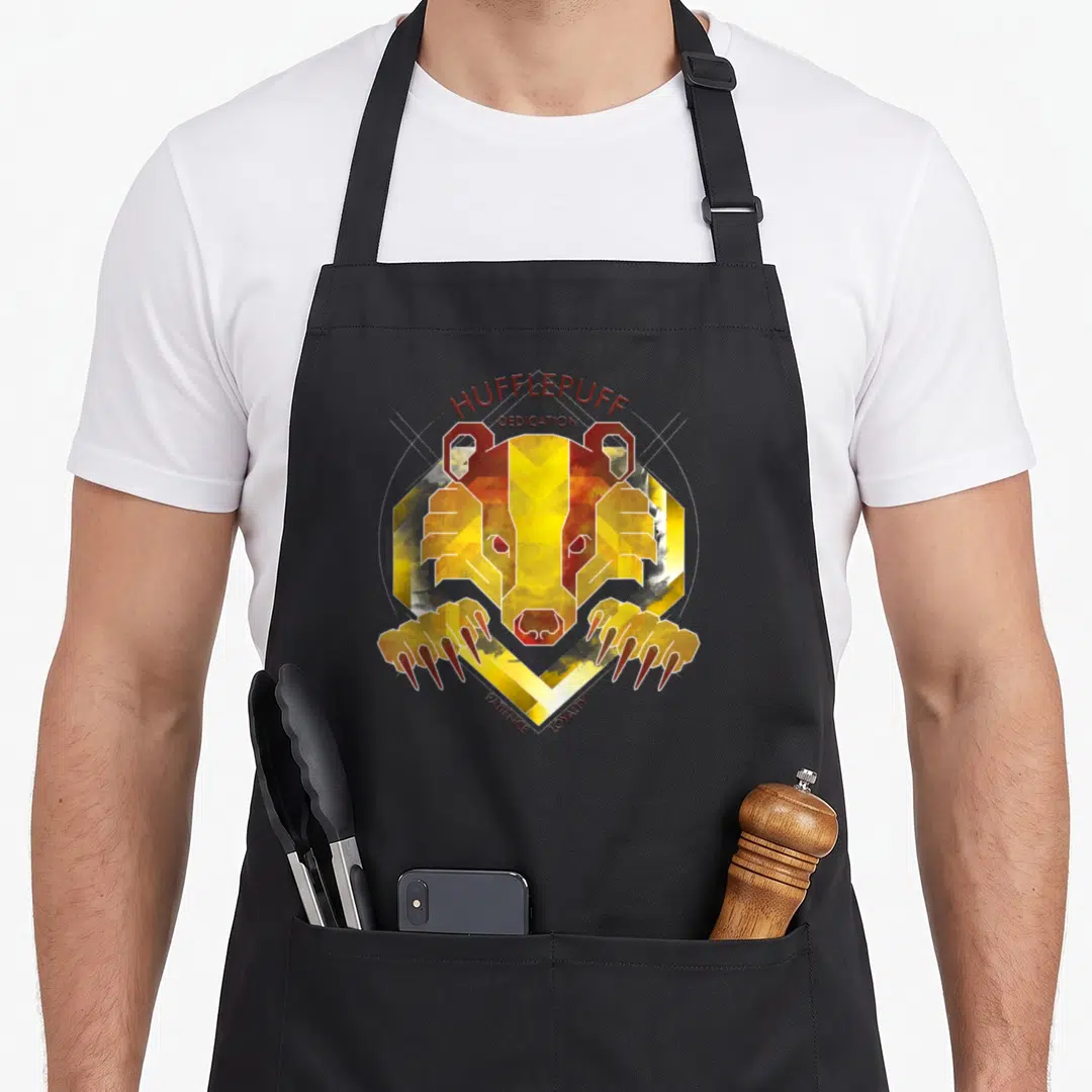 Dedication hufflepuff apron - Geometric badger head with claws and text Hufflepuff Dedication Patience Loyalty — shown on a black cotton canvas apron against a white background