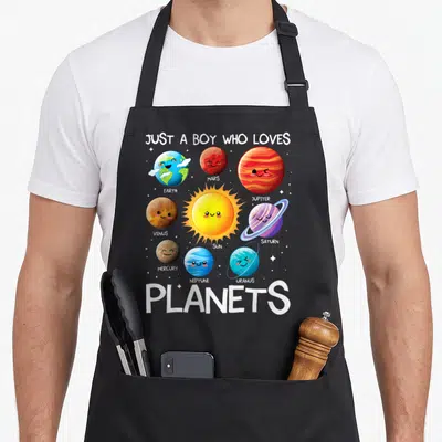 Planet solar science apron - Cartoon solar system with labeled planets and central sun under Just A Boy Who Loves — shown on a black cotton canvas apron against a white background