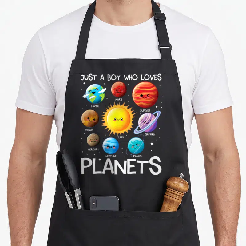 Planet solar science apron - Cartoon solar system with labeled planets and central sun under Just A Boy Who Loves — shown on a black cotton canvas apron against a white background