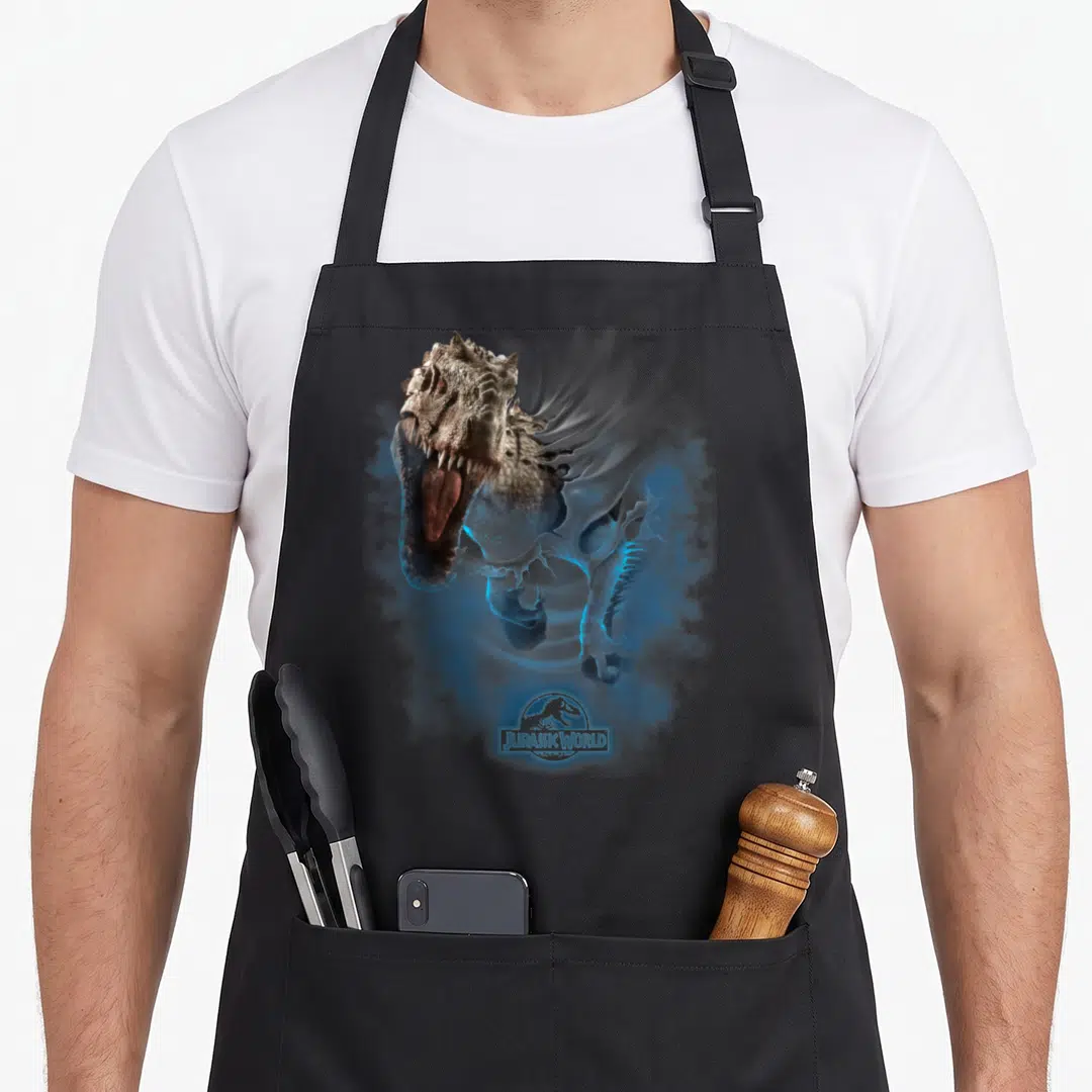 Through t-rex apron - Indominus Rex head dissolving into blue smoke above Jurassic World logo — shown on a black cotton canvas apron against a white background